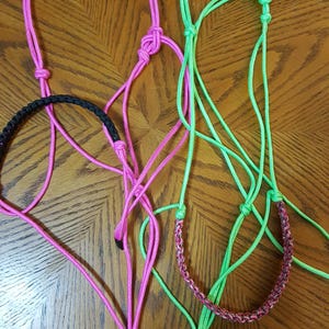 May include: Three rope trail halters in pink, green, and black are displayed. The pink halter has a matching lead rope. The green halter has a braided noseband. The halters are on a wooden surface. The tag reads "DO NOT USE TO TEACH TYING".