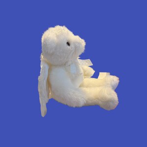 May include: A white plush bunny toy with long ears and a small bow. The bunny is sitting down, with its legs extended. The toy is made of soft, fluffy material and has black eyes.
