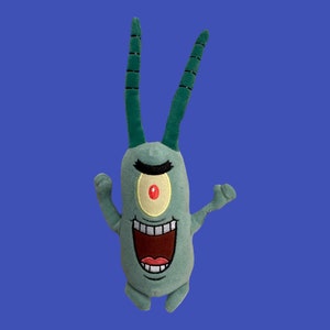 May include: A green plush toy of a single-celled organism with a large red eye and a wide, toothy grin. The toy is in a playful pose with its arms raised.