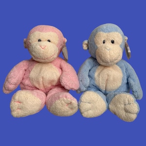 以下が含まれることがあります： ピンクとブルーのぬいぐるみのおさるさんの玩具が2体、並んで座っています。両方のサルのお腹と顔は白い毛皮です。