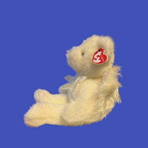 May include: A cream-colored plush teddy bear with a white bow and a red heart-shaped tag. The bear is sitting on a blue background. The tag reads "Classic".