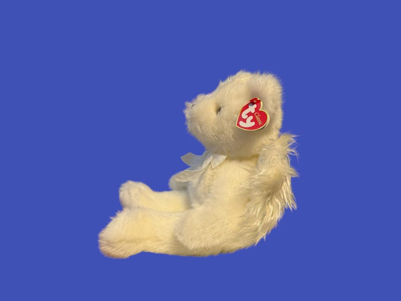 May include: A cream-colored plush teddy bear with a white bow and a red heart-shaped tag. The bear is sitting on a blue background. The tag reads "Classic".