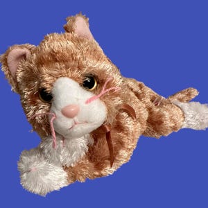 Op de afbeelding: Een pluche oranje en witte katten speelgoed met roze gestikte snorharen en bruine ogen. De kat ligt op zijn zij met zijn hoofd gedraaid.