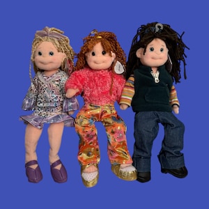 May include: Three plush dolls with different hairstyles and outfits. The doll on the left has blonde hair and wears a patterned dress and purple shoes. The middle doll has red hair and a pink top with floral pants. The doll on the right has dark hair, a green vest, and jeans.