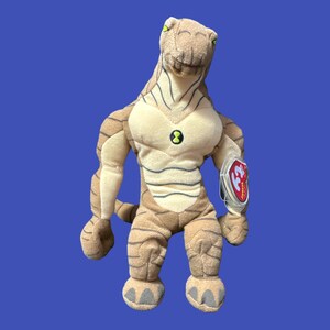 K&ouml;nnte beinhalten: Ein braunes und beiges Pl&uuml;schtier eines humanoiden Au&szlig;erirdischen mit einem gr&uuml;nen Symbol auf der Brust. Das Spielzeug hat ein rotes Beanie Baby-Etikett am Arm.