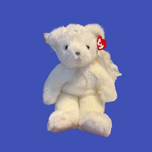 May include: A white plush teddy bear with angel wings and a bow. The bear has a red tag attached to its ear. The bear is sitting on a blue background.