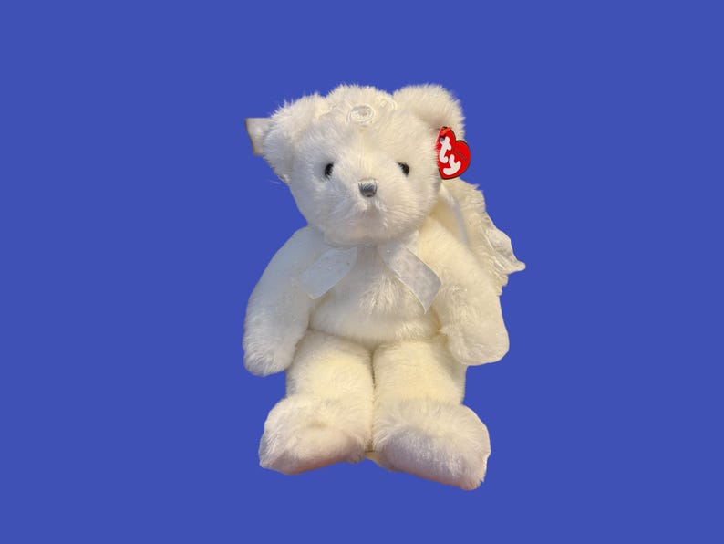 May include: A white plush teddy bear with angel wings and a bow. The bear has a red tag attached to its ear. The bear is sitting on a blue background.