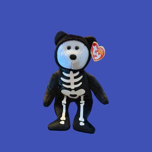 May include: A plush bear toy featuring a black body with a white skeleton print on the front and a white face. The bear has a black hood and a heart-shaped red and yellow Ty tag attached to its ear, displaying the text Ty and Beanie Baby.