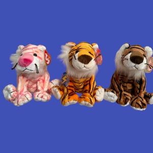 Könnte beinhalten: Drei Plüschtiger. Einer ist pink und weiß, einer ist orange und schwarz und einer ist braun und schwarz.