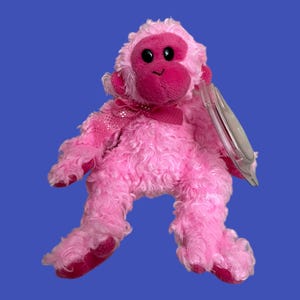 Peut inclure: Singe en peluche rose avec un nœud rose pailleté et une étiquette en plastique transparente. Le singe a un visage rose avec des yeux noirs et un nez rose.