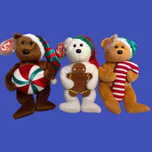May include: Three Beanie Baby bears dressed for the holidays. A brown bear holds a peppermint candy, a white bear holds a gingerbread man, and a tan bear wears a striped scarf. Each bear has a tag attached.