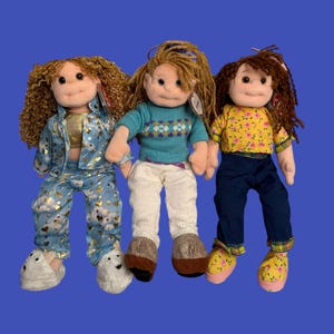 Peut inclure: Trois poupées en peluche aux tenues variées. L'une porte un pyjama bleu avec des accents dorés, une autre un pull sarcelle et un pantalon blanc, et la troisième un haut jaune à fleurs et un pantalon marine. Chaque poupée a une coiffure unique.