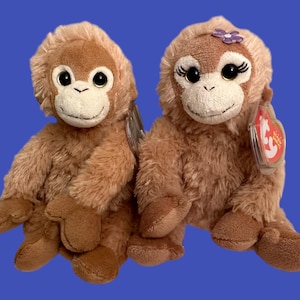 以下が含まれることがあります： 茶色のぬいぐるみのおさるのビーニーベイビーズが2体。1体のおさるは紫色の花を頭に付けています。2体とも赤いビーニーベイビーズのタグが付いています。