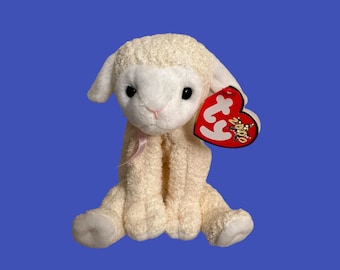 May include: A white plush lamb with a pink ribbon around its neck. The lamb is sitting with its legs crossed. It has a red Ty tag with the year 2000 on it.