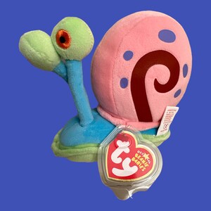 May include: A pink and blue snail plush toy with a red swirl on its shell. The toy has a Ty Beanie Babies tag attached to it.