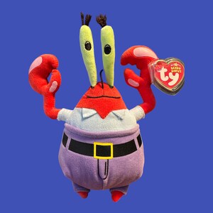 May include: A red and purple plush toy crab wearing a blue shirt and a black belt. The crab has green eyes and a red heart-shaped tag that says "Beanie Babies" and "Ty".