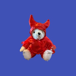 Pode incluir: Um urso de peluche branco vestido com um traje de diabo de cetim vermelho brilhante. O traje inclui uma capa com capuz com dois chifres pontiagudos no topo e uma fita no pescoço. O urso tem olhos e nariz pretos, com uma etiqueta vermelha.