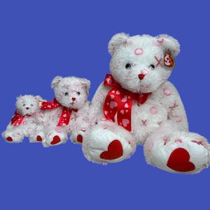 May include: Three white plush teddy bears with red hearts and pink hearts printed on their fur. The bears are wearing red and pink ribbon bows around their necks. The largest bear is sitting with its legs outstretched. The two smaller bears are sitting behind the larger bear.