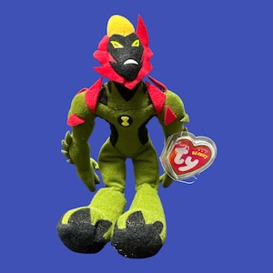 K&ouml;nnte beinhalten: Gr&uuml;nes und rotes Pl&uuml;schtier eines Charakters mit einem Ty Beanie Babies-Etikett. Der Charakter hat einen gro&szlig;en Kopf mit einem roten und schwarzen Gesicht und einem gelben Kamm. Der Charakter hat einen gr&uuml;nen K&ouml;rper mit schwarzen F&uuml;&szlig;en und H&auml;nden.