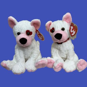 May include: Two white plush dogs with pink noses and red hearts on their eyes. The dogs are sitting side by side and have red and white Ty beanie tags attached to their necks.