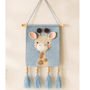May include: A handmade crochet wall hanging featuring a giraffe design. The giraffe has a beige face, brown spots, and pink ears, set against a light blue background. The piece is finished with blue tassels and wooden beads. The text "Maple & Moon Tales" is at the bottom.