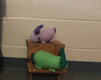 May include: Two crocheted stuffed animals, a purple bunny and a green worm, sitting on a small wooden stool.