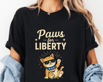 May include: Black t-shirt with the text "Paws for Liberty" and a cartoon cat wearing sunglasses and a patriotic bandana. The cat is waving. The shirt is a casual style.