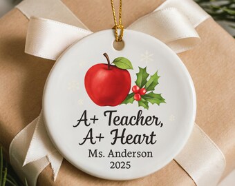 May include: White ceramic ornament with a red apple and holly design. The ornament has the text "A+ Teacher, A+ Heart" and "Ms. Anderson 2025". A gold string is attached for hanging. The ornament is on a gift box with a cream ribbon.