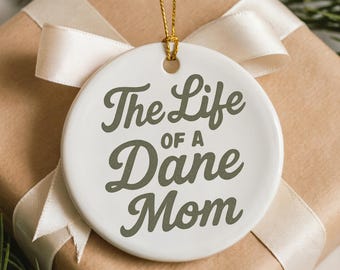 May include: A white ceramic ornament with the text "The Life of a Dane Mom" in a dark green script. The ornament hangs from a gold string and is set against a wrapped gift with a cream-colored ribbon.
