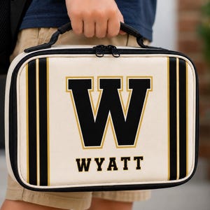 May include: A rectangular lunch bag in cream with black and gold vertical stripes on the sides. The front features a large black letter W with a gold outline, and the name WYATT printed in black capital letters underneath. It has a black handle.