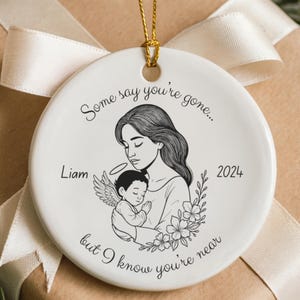 Può includere: Ornamento natalizio in ceramica bianca con cordino dorato. Presenta un'illustrazione in bianco e nero di una madre e un bambino, con il testo "Some say you're gone..." e "but I know you're near."