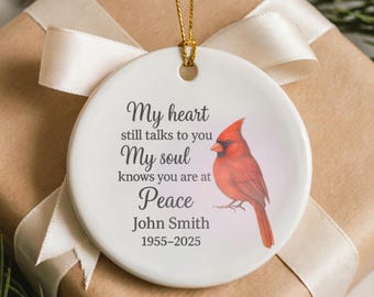 May include: A white ceramic ornament with a red cardinal illustration and the text "My heart still talks to you, My soul knows you are at Peace, John Smith 1955-2025." The ornament hangs from a gold string and is set on a gift box with a cream ribbon.