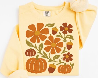 May include: A pale yellow crewneck sweatshirt featuring a graphic print of orange flowers, green leaves, brown acorns, and orange pumpkins. The inner neck label displays the text Comfort Colors. The long-sleeved garment has a ribbed collar.