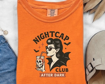 May include: Orange t-shirt with a graphic featuring a woman in sunglasses drinking from a skull-shaped glass, with the words "NIGHTCAP CLUB AFTER DARK". The shirt has a crew neck and short sleeves.