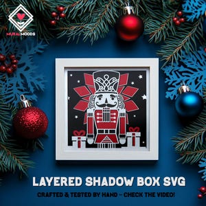 May include: A framed layered shadow box featuring a red and white nutcracker design on a black background. The frame is white, and the image is surrounded by Christmas ornaments and evergreen branches. Text at the bottom reads "LAYERED SHADOW BOX SVG".