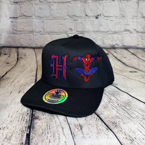 Op de afbeelding: Een zwarte snapback-pet met een geborduurde rode en blauwe letter H en een rood en blauw Spider-Man-figuur. Een ronde holografische sticker op de klep toont de tekst FROG PRINTS ORIGINAL SNAPBACK.