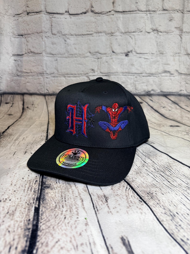 Op de afbeelding: Een zwarte snapback-pet met een geborduurde rode en blauwe letter H en een rood en blauw Spider-Man-figuur. Een ronde holografische sticker op de klep toont de tekst FROG PRINTS ORIGINAL SNAPBACK.