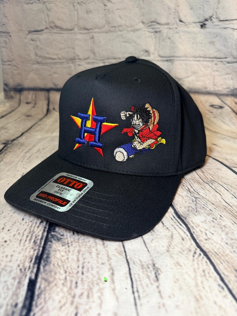 Op de afbeelding: Een zwarte honkbalpet met een geborduurde blauwe letter H over een rode en gele ster aan de linkerkant en een kleurrijk anime-personage met een strooien hoed aan de rechterkant. Een zilveren sticker op de klep vermeldt OTTO CLASSIC FIT OSFM MID PROFILE.