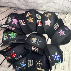 May include: A collection of black baseball caps featuring embroidered designs of various animated characters and a stylized letter H. Each cap includes a prominent Otto brand sticker on the brim, showcasing diverse color palettes and graphic themes.