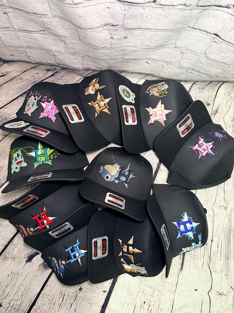 Op de afbeelding: Een verzameling zwarte baseballpetten met geborduurde ontwerpen van diverse geanimeerde figuren en een gestileerde letter H. Elke pet is voorzien van een opvallende Otto-sticker op de klep en toont uiteenlopende kleurenpaletten.