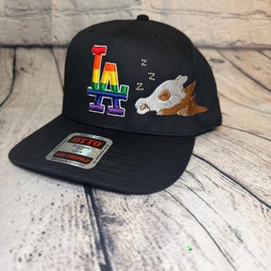 Op de afbeelding: Een zwarte honkbalpet met een geborduurd regenboogkleurig LA-logo en een slapend wezen met een schedelmasker en bruin lichaam. Drie witte Z's zijn erboven geborduurd. Een Otto-sticker bevindt zich op de platte klep van de pet.