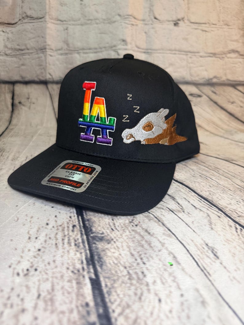 Op de afbeelding: Een zwarte honkbalpet met een geborduurd regenboogkleurig LA-logo en een slapend wezen met een schedelmasker en bruin lichaam. Drie witte Z's zijn erboven geborduurd. Een Otto-sticker bevindt zich op de platte klep van de pet.