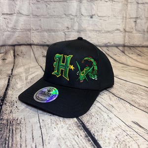 Op de afbeelding: Een zwarte snapback-pet met een geborduurde groene en gele letter H naast een groen draakachtig wezen. Een ronde holografische sticker op de klep toont de tekst Frog Prints Original Snapback in zwarte en witte letters.