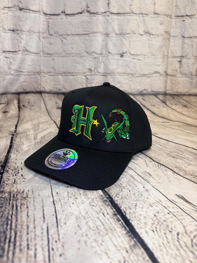 Op de afbeelding: Een zwarte snapback-pet met een geborduurde groene en gele letter H naast een groen draakachtig wezen. Een ronde holografische sticker op de klep toont de tekst Frog Prints Original Snapback in zwarte en witte letters.