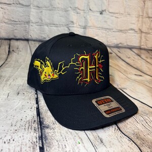 Op de afbeelding: Een zwarte baseballpet met een geborduurde gele Pikachu naast een gestileerde letter H in rood en geel met bliksemschichten. Een sticker op de klep bevat de tekst Otto Classic Fit Mid Profile Premium Fabric.