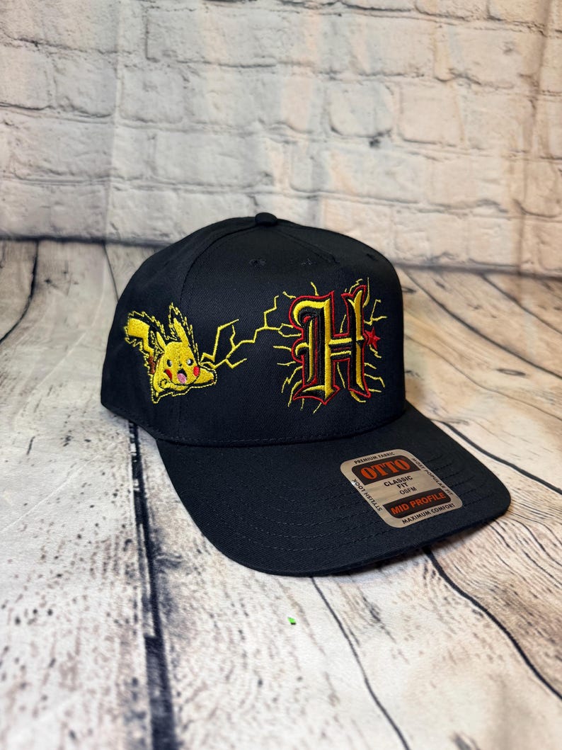 Op de afbeelding: Een zwarte baseballpet met een geborduurde gele Pikachu naast een gestileerde letter H in rood en geel met bliksemschichten. Een sticker op de klep bevat de tekst Otto Classic Fit Mid Profile Premium Fabric.