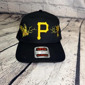 Op de afbeelding: Een zwarte honkbalpet met een gele geborduurde letter P in het midden, geflankeerd door gele geborduurde Pikachu- en Zapdos-figuren met bliksemschichten. Een zilveren en zwarte sticker op de klep vermeldt OTTO CLASSIC FIT OSFM.