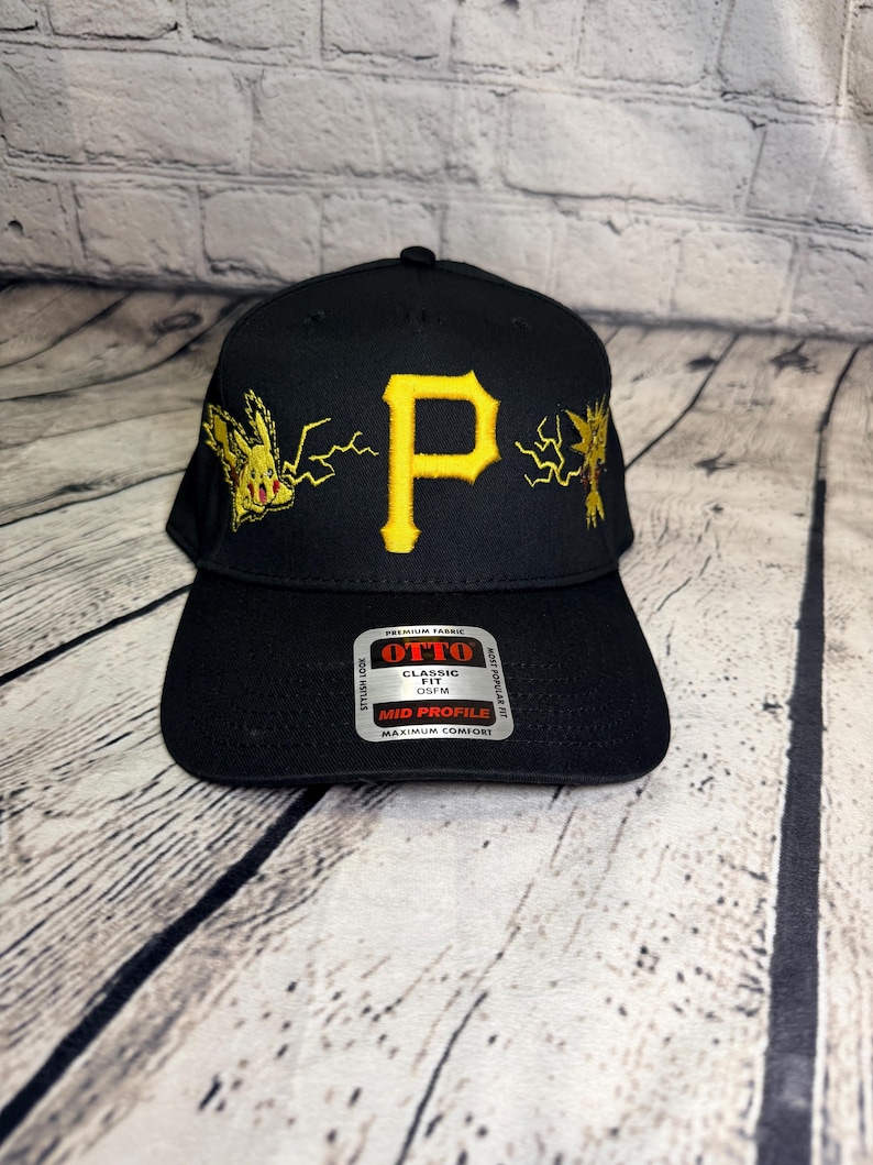 Op de afbeelding: Een zwarte honkbalpet met een gele geborduurde letter P in het midden, geflankeerd door gele geborduurde Pikachu- en Zapdos-figuren met bliksemschichten. Een zilveren en zwarte sticker op de klep vermeldt OTTO CLASSIC FIT OSFM.