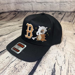 Op de afbeelding: Een zwarte honkbalpet met een geborduurde gouden letter B en een wit en bruin stripfiguur dat een bot vasthoudt. De pet heeft een sticker op de klep met de tekst OTTO, CLASSIC FIT, OSFM, MID PROFILE en MAXIMUM COMFORT.