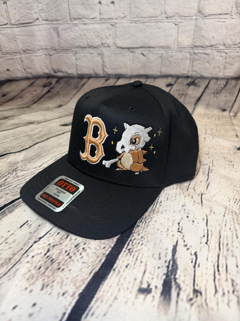 Op de afbeelding: Een zwarte honkbalpet met een geborduurde gouden letter B en een wit en bruin stripfiguur dat een bot vasthoudt. De pet heeft een sticker op de klep met de tekst OTTO, CLASSIC FIT, OSFM, MID PROFILE en MAXIMUM COMFORT.